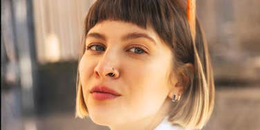 Woman with a brown bob and bangs, a nose ring, and orange sunglasses on her head looking at the camera with a calm, confident expression.