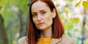 Red-haired woman in a mustard sweater and tan coat looking off-camera with a hurt, guarded expression in a park.