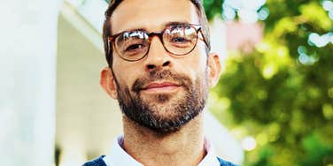 Bearded man in tortoiseshell glasses and a blue sweater looking at the camera with a slight smirk outdoors