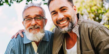 Older man with a white beard and glasses smiling beside an adult man with a beard, who has an arm around his shoulder, outdoors