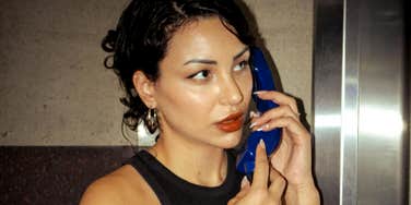 A close-up portrait of a dark-haired woman with hoop earrings holding a blue vintage telephone receiver to her ear, glancing sideways with a calm, expectant expression, representing casual offloading of effort and confident entitlement.
