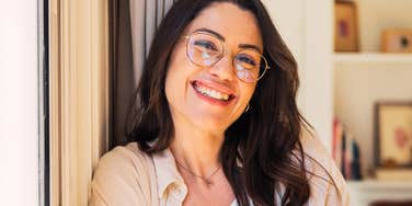 People Who Like To Sound Smart Often Say Phrases Without Realizing What They're Even Saying A dark-haired woman in stylish round glasses leaning against a doorway and laughing warmly, symbolizing approachable intelligence, clear speech, authentic personality, and relaxed conversation.