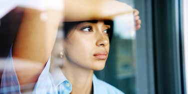 A close-up shot through glass of a young woman with dark hair and bangs resting her hand against her forehead while looking out a window with a serious, pensive expression, representing environmental reflection, isolation, and energy flow.