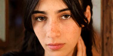 Young woman with dark hair and freckles looking directly at the camera with a guarded, unreadable expression, one hand at her neck.