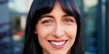 A close-up portrait of a brunette woman with dark bangs and hazel eyes wearing mauve lipstick, smiling confidently and warmly into the camera outdoors, representing self-assurance, emotional balance, and natural charisma.