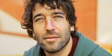 Man with curly dark hair, light eyes, and small hoop earrings looking calmly at the camera in a green hoodie against an orange background