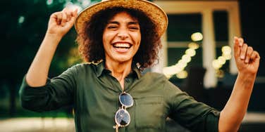 Smiling woman in a straw hat and green shirt laughing with her arms raised, string lights glowing behind her.