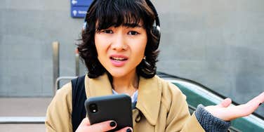 Woman with dark short wavy hair, wearing headphones and holding their phone while looking up with a confused, questioning face. 