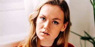 Woman with long blonde hair and a red sweater looking directly at the camera beside a window and a leafy plant