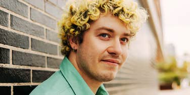 Young man with curly blond hair and a mint green shirt leaning against a dark brick wall, looking off to the side with a slight smile