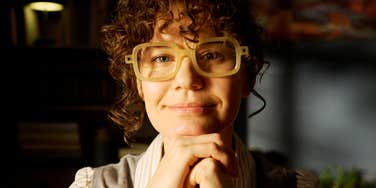 A close-up portrait of a woman with curly brunette hair and thick yellow-framed glasses resting her chin on her hands in a warm, dimly lit room, smiling with calm confidence and intellect.