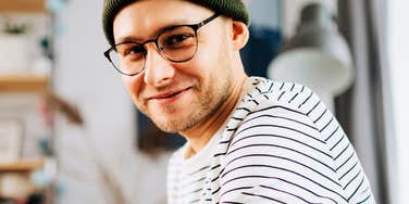 Close-up portrait of a cheerful young man wearing black-rimmed glasses, a green beanie, and a striped shirt smiling casually in his living room.