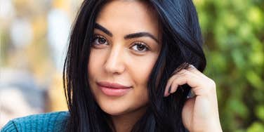 A close-up portrait of a woman with long dark hair and brown eyes looking directly at the camera with a subtle smile while gently touching her hair, representing strong eye contact, magnetic presence, and interpersonal warmth.