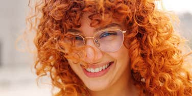 Smiling woman with curly red hair and thin gold glasses tilting her head toward the camera