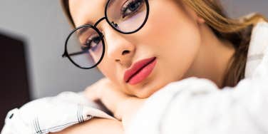 A close-up portrait of a young blonde woman wearing stylish round black glasses and a white button-down shirt, resting her head on her crossed arms with a weary, contemplative expression, representing workplace fatigue, overwork, and professional boundary struggles.
