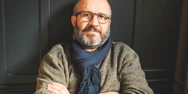 A middle-aged man with a gray beard and glasses wearing a olive green sweater and dark blue scarf sitting with crossed arms against a dark backdrop, looking thoughtfully into the camera, representing healthy aging, wisdom, and cognitive focus.