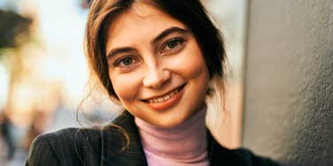 Portrait of a frugal brunette woman wearing a casual blazer, leaning on a wall outside. 
