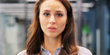 Woman with long brown hair in a blue striped shirt looking worried and confused at the camera in a blurred office setting.