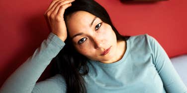 Woman with long dark hair and freckles resting her head on her hand and looking tired and unhappy at the camera against a red wall.