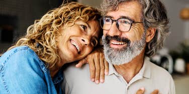 Smiling middle-aged couple at home, the curly-haired woman resting her head and hand on her bearded husband’s shoulder as they look off-camera.