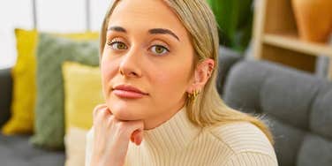 A blonde woman in a cream turtleneck sitting on a gray sofa resting her chin on her hand, looking into the camera with a detached, emotionless expression, representing marital burnout, emotional disconnection, and indifference.