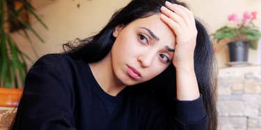 Woman with long dark hair resting her hand on her forehead and looking tired and emotionally drained at the camera in a warmly lit room.