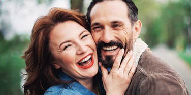 Laughing middle-aged couple pressed cheek to cheek outdoors, the woman with red hair and red lipstick, the man with a dark beard, hugging in a park.