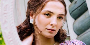 A young woman with soft brown waves and white rose earrings looking calmly into a framed mirror outdoors against a log cabin wall, representing self-reflection, personal value, poised confidence, and boundary setting.