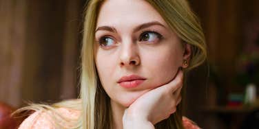 A close-up portrait of a young blonde woman with hazel eyes wearing a light peach top, resting her chin on her hand while looking off to the side with a tired, contemplative expression, representing sleep deprivation, fatigue, and female health challenges.