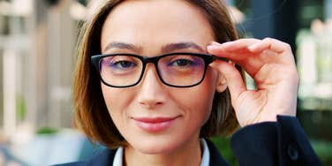 Woman with a short brown bob adjusting black-framed glasses and smiling confidently at the camera, wearing a navy blazer.