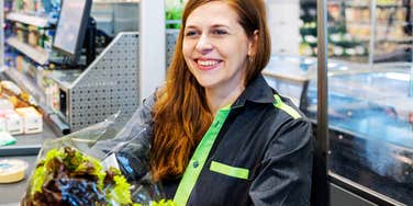 A friendly female grocery store cashier with long reddish-brown hair in a gray and neon green uniform smiling brightly while scanning fresh salad greens at a checkout counter, representing frontline patience, customer service, and human observation.