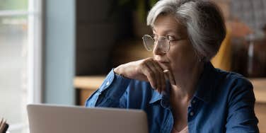 older woman looking out window with laptop in front of her sitting at table
