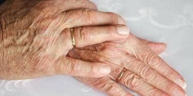 An elderly couple holds hands with wedding bands on their fingers.