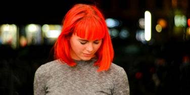 young white woman with bright red dyed hair in a beige sweater looks down, seemingly in pain