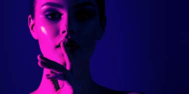 woman in pink and purple lighting putting her finger to her lips to shush