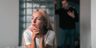 woman looking away sitting on bed husband blurred in background