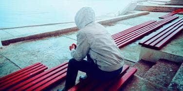 man in hoodie sitting, depressed, on a park bench looking at ocean