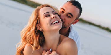 A tanned young caucasian bearded guy kisses a beautiful fun smiling blonde woman against the backdrop of the setting sun.