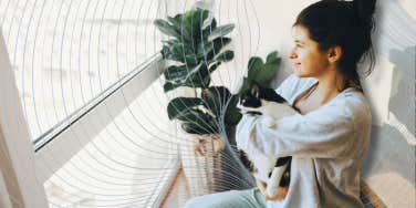 Sensitive woman holding her cat looking out window 