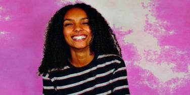 young woman with dark skin and black hair smiles broadly in front of pink background