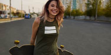 Cheerful girl with tattoos posing with a longboard while walking on the road in the city