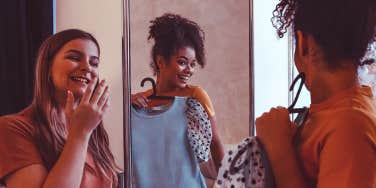 A photograph of two women looking into a mirror, with one holding up a blouse to herself with a grin.