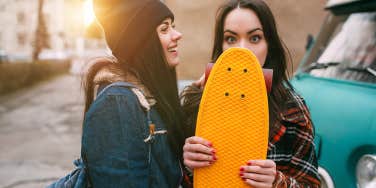 Two street girls smiling and joking with skateboards near vintage cars