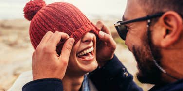 Loving couple having fun outdoors. Man covering eyes of woman with cap.