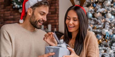 spouse giving Christmas gift to his girlfriend sitting beside a Christmas tree at home.