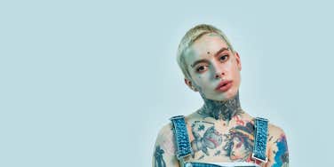  tattooed and pierced white girl wearing denim overall standing and looking into a camera on a light blue color background
