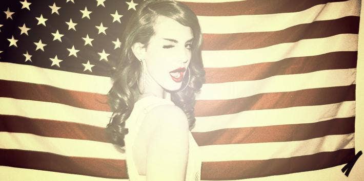 Lana Del Rey winking wearing red lipstick in front of the United States American flag for her "Ride" music video