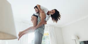 Young man standing on bed and carrying his girlfriend on his shoulder. Couple in playful mood in bedroom.