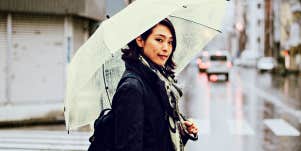 Asian woman crossing rainy street with umbrella, looking sweetly at camera