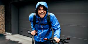 white guy with curly hair laughs into camera, wearing blue rain coat holding a mountain bike